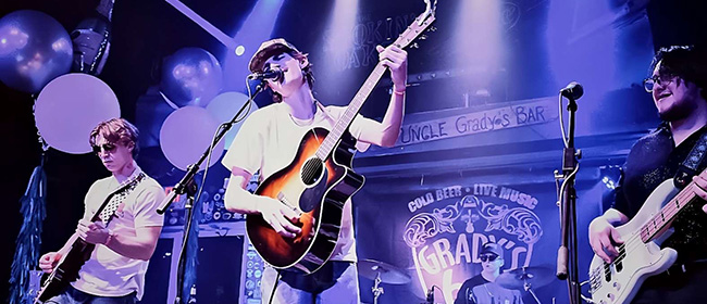 Three band members on a stage, with the main signer playing an acoustic guitar