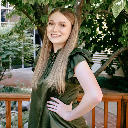 Woman smiles while standing in front of a wooden railing and leafy green trees