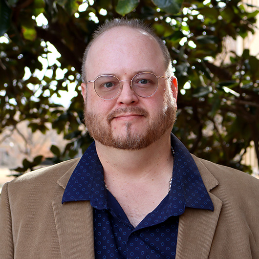 Man wth round glasses and a might brown beard. Wearing a blue shirt with blue dots, and a tan corduroy jacket.