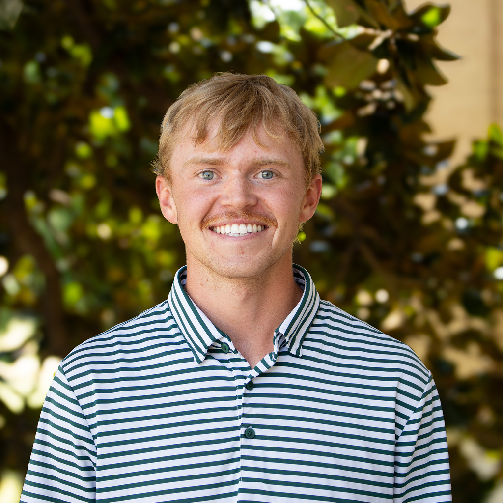 A man in striped button-up shirt standing and smiling in front of a tree
