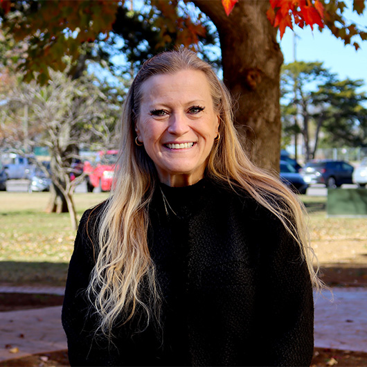 Woman with long blonde hair and a black sweater stands in front of a tree with bright red fall foliage