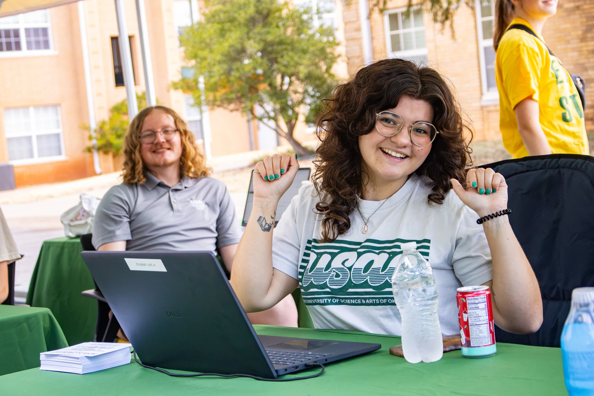 Smiling USAO student raises her hands while seated at a table with a laptop during an outdoor campus event, with another student in the background.