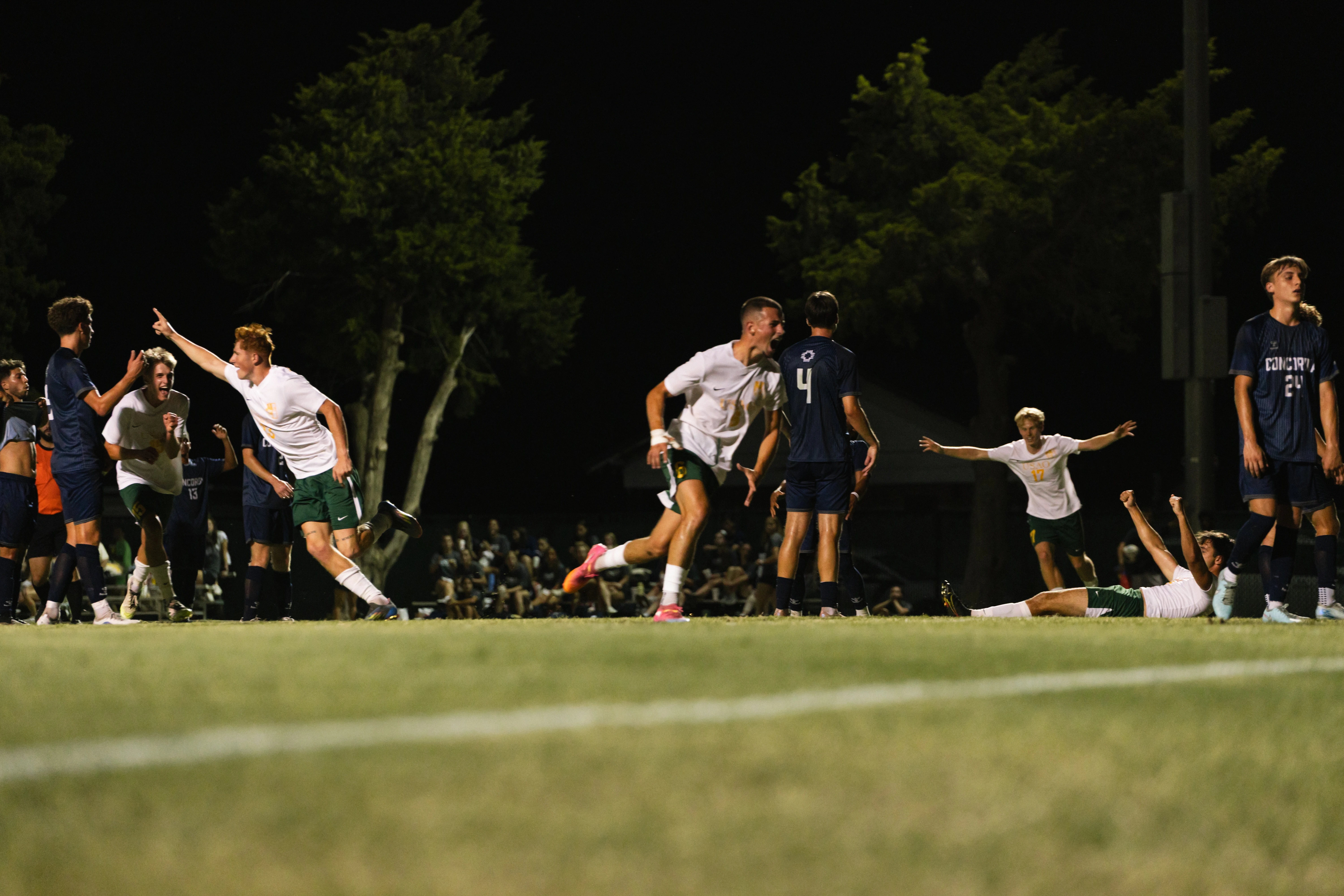 USAO men’s soccer players celebrate during a nighttime match as opposing players stand nearby.